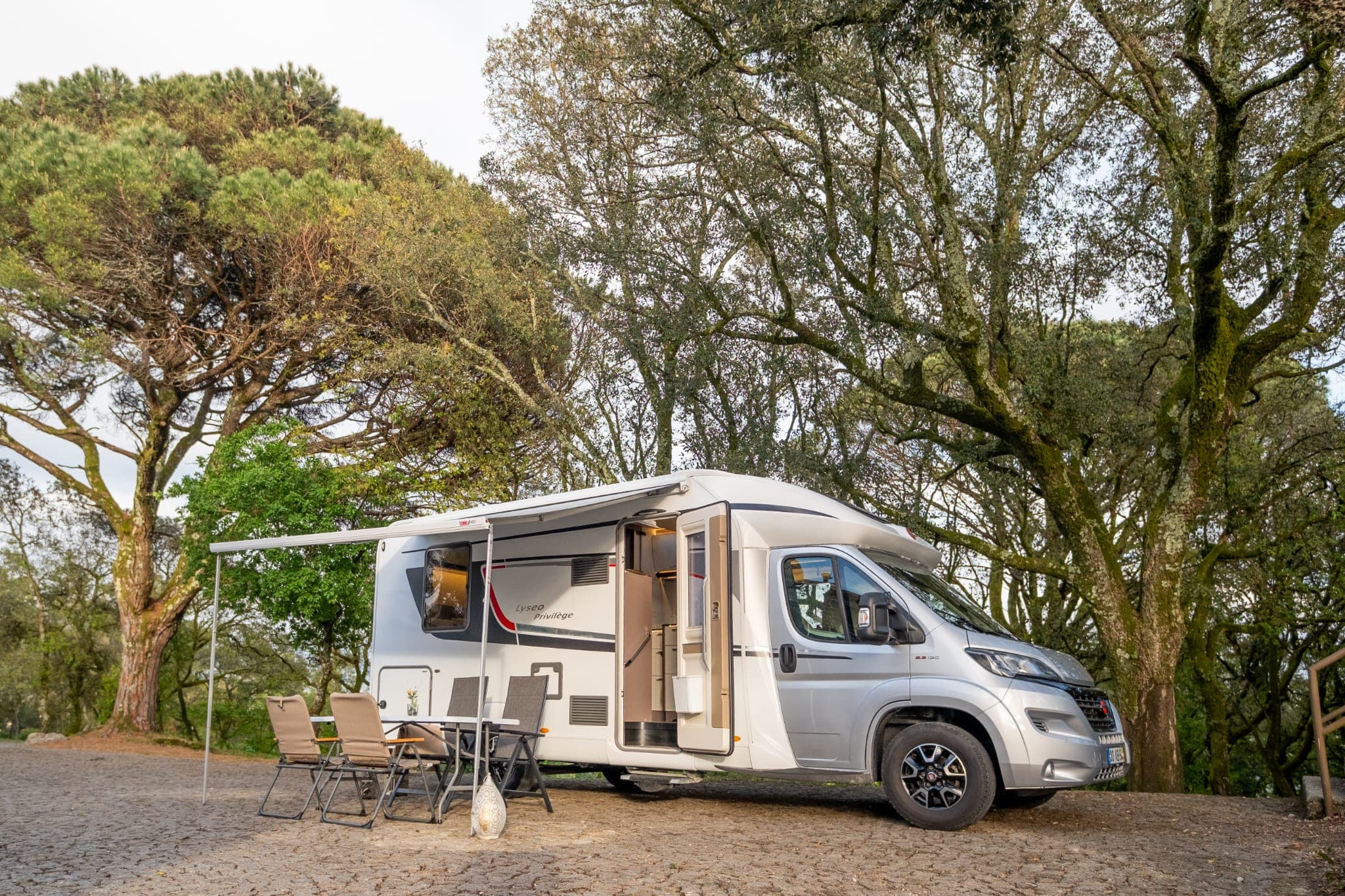 Autocaravana estacionada entre árvores, com toldo aberto e mesa de exterior